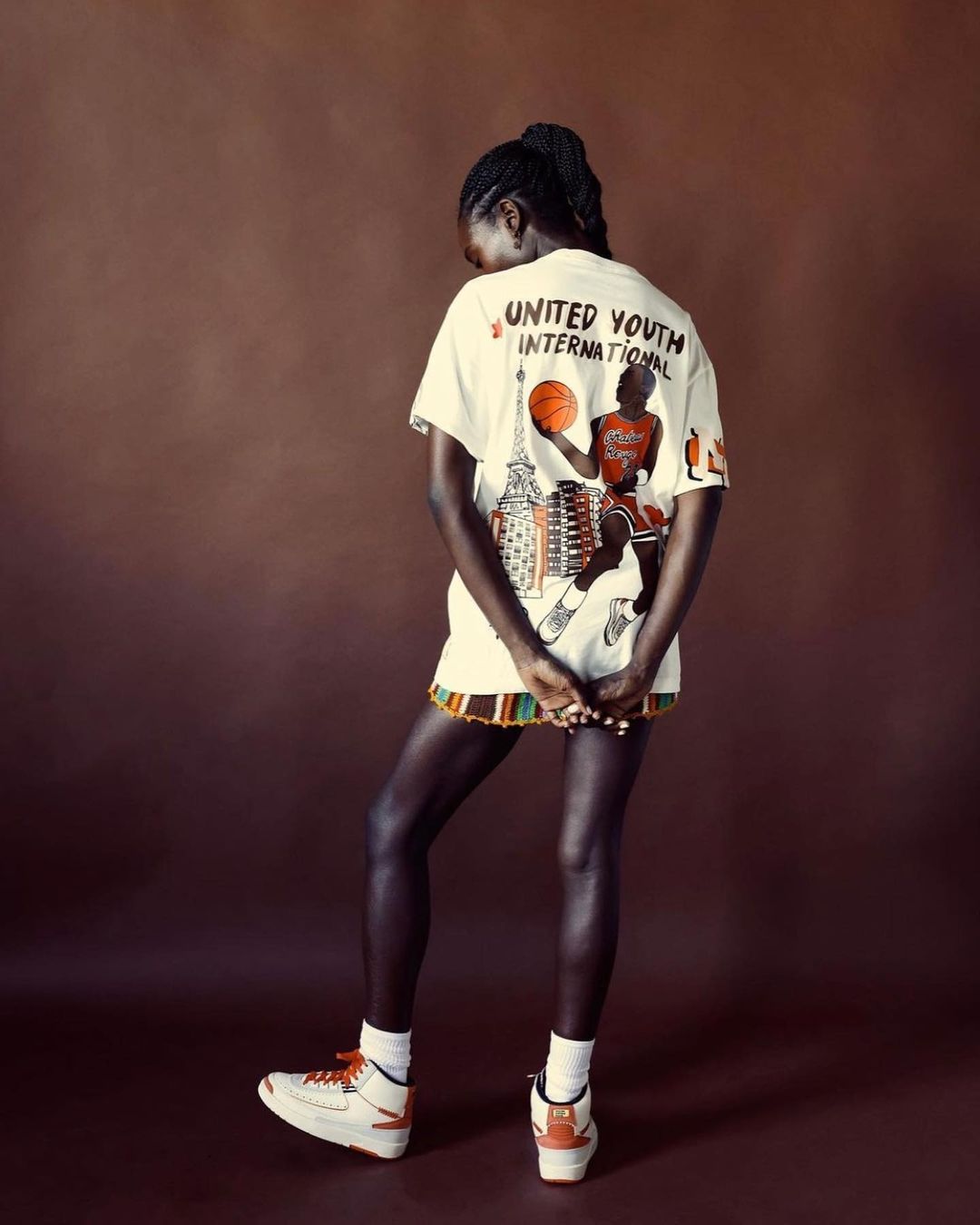 Standing Black woman in t-shirt dress with graphic, sneakers, and high socks against a solid brown background in a studio shot.