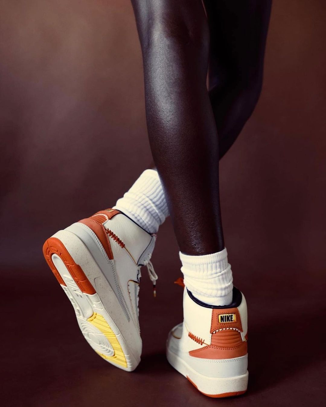 A person stands in high-top Nike sneakers and white socks, showcasing the footwear's design against a solid brown background.