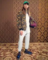 Young man with long hair wearing a patterned jacket, white shirt, grey sweatpants, green cap, and blue loafers standing indoors.