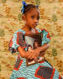Young Black girl holding a framed photograph while wearing a patterned dress against a patterned wallpaper background