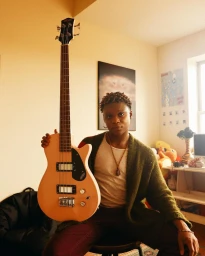 Black man holding up a light orange bass guitar in a brightly lit room, photographed by Lawrence Agyei