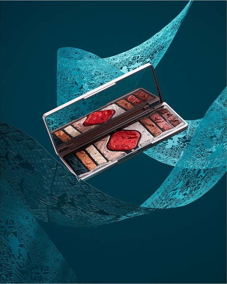 Eye shadow palette with multiple colors and a mirror, presented on a teal backdrop with decorative swirls for a beauty product shot.