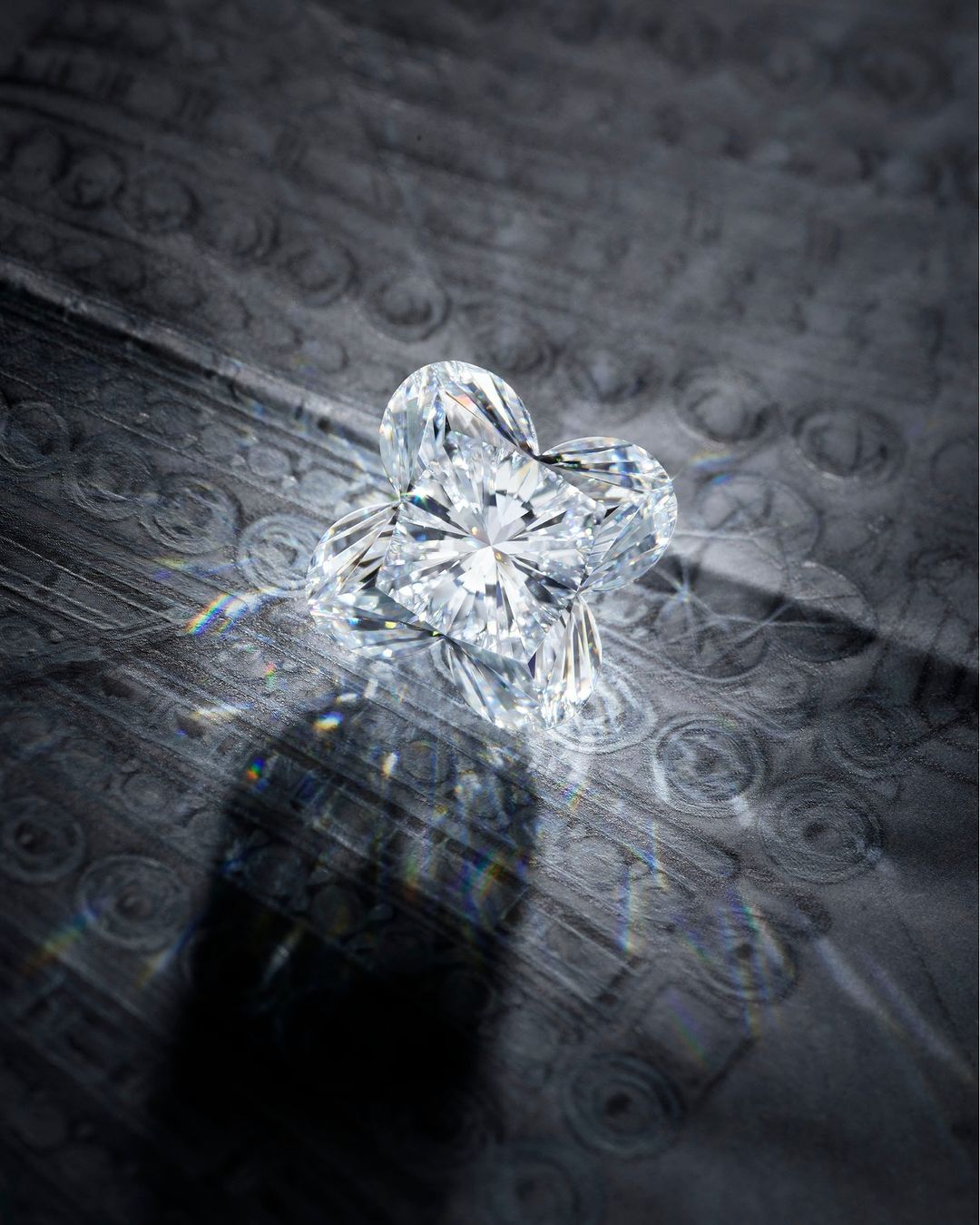 Heart shaped diamond with facets sits on a patterned dark grey reflective surface, casting a dark shadow and refracting light.