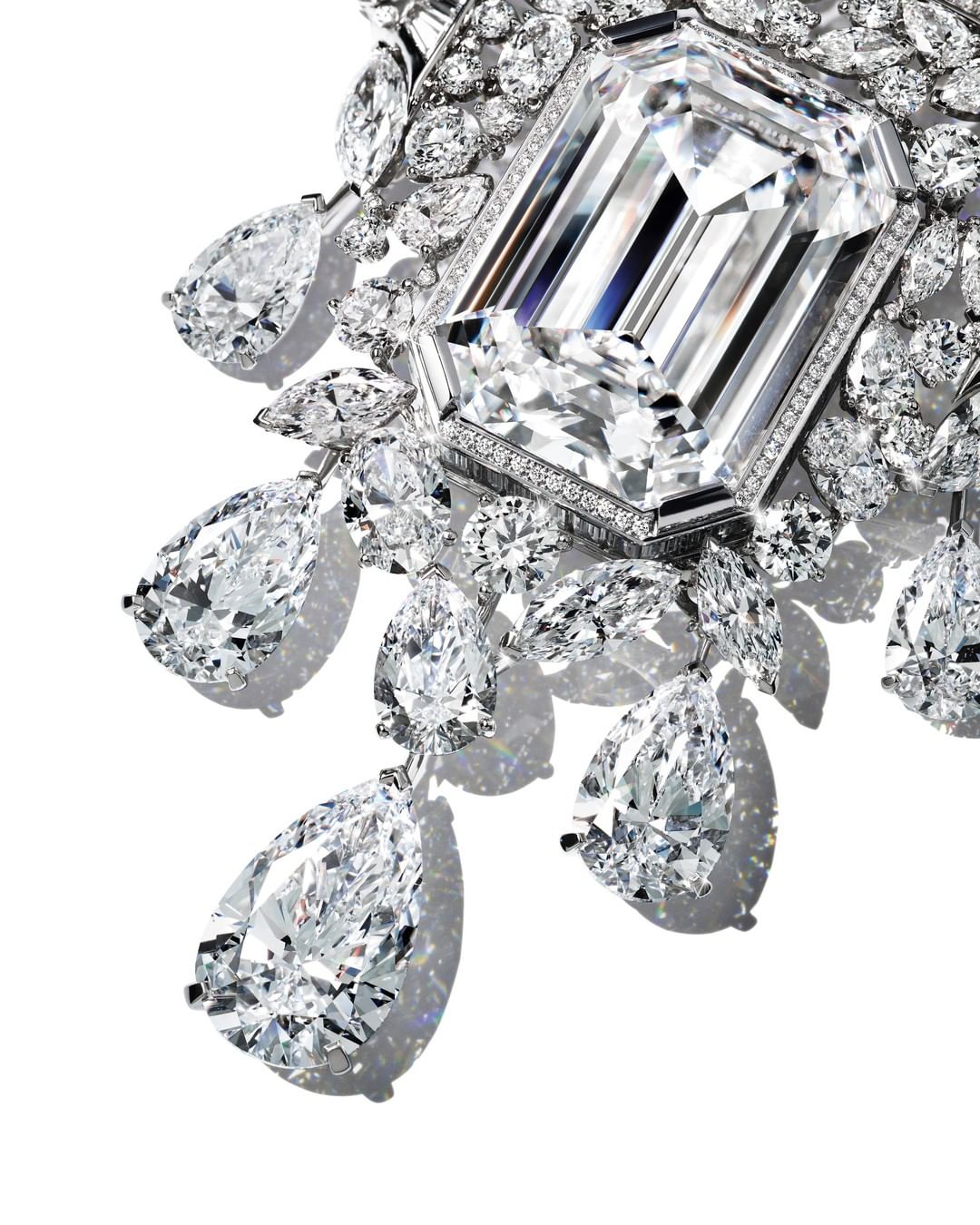 Close-up of an ornate diamond necklace featuring a large emerald-cut diamond surrounded by smaller, pear-shaped diamonds on a white background.