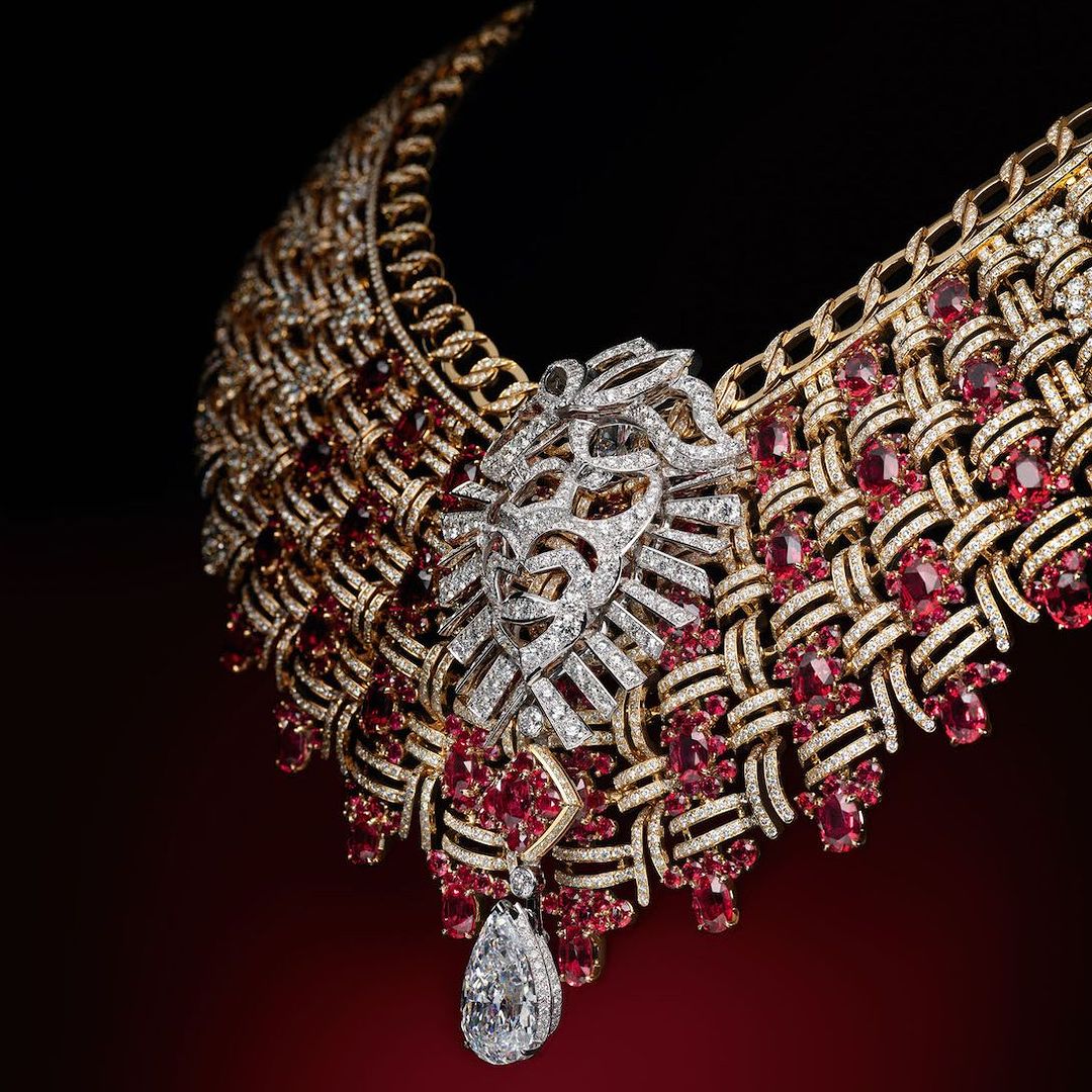 Close-up of a gold statement necklace with white diamond and ruby gemstones in a repeating woven pattern. Red and black background.