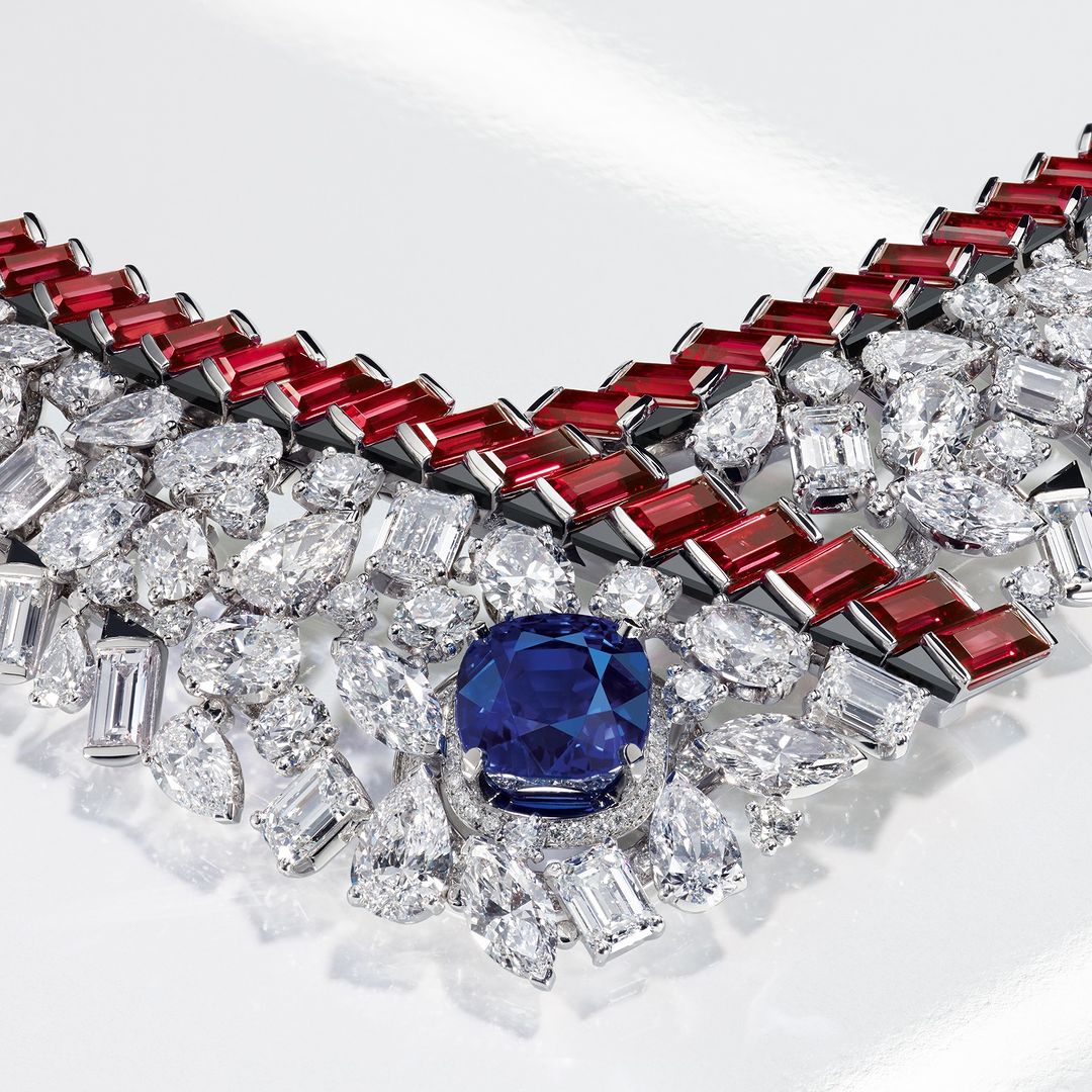 Close-up of a V-shaped statement necklace featuring large blue, white, and red gemstones set in a light-reflecting metal, against a white backdrop.