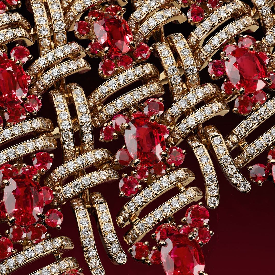 Close-up of a woven gold necklace featuring ruby and diamond floral accents, set against a dark red background. Jewelry advertisement.