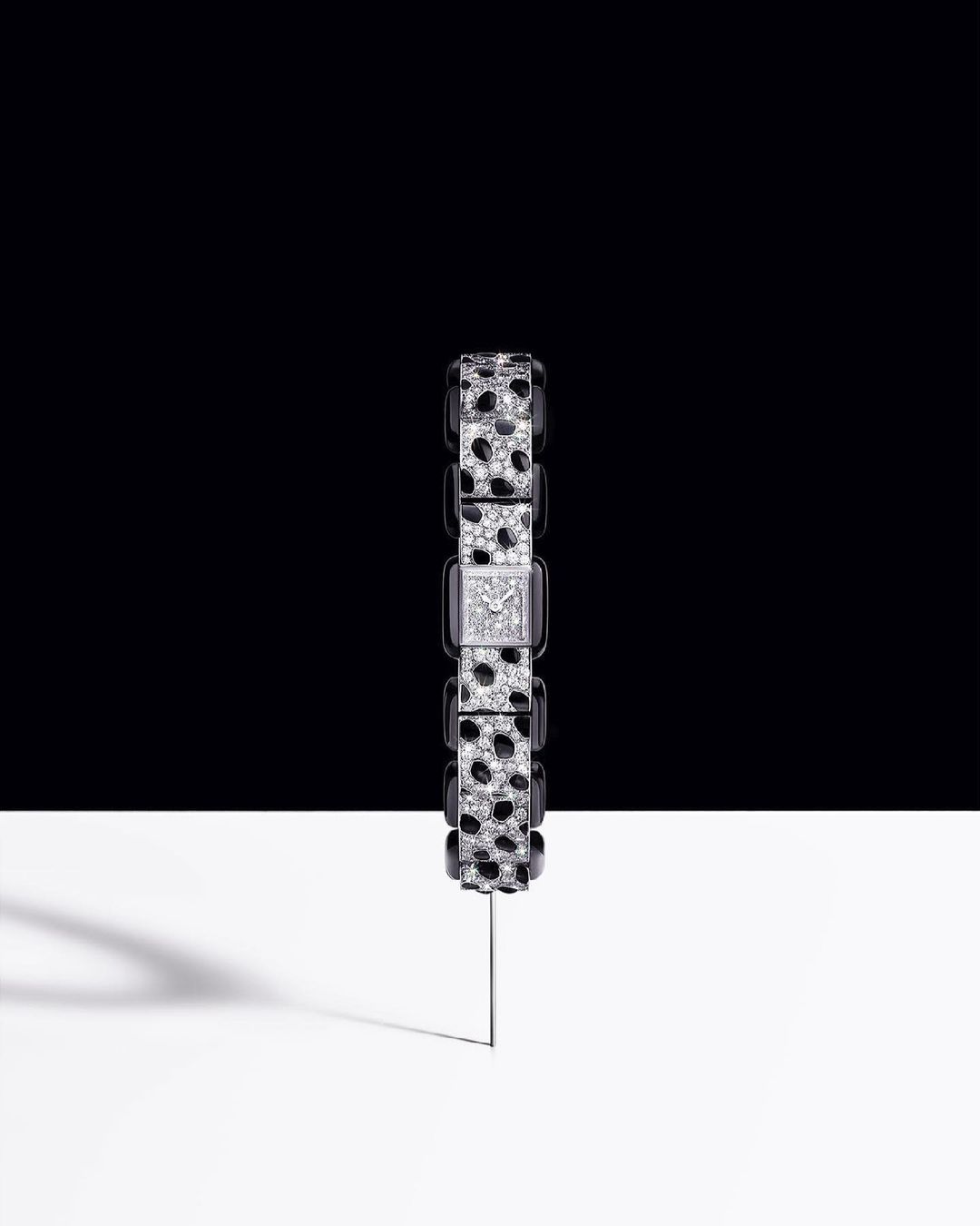 A black and white wristwatch with gemstones is displayed against a split black and white background, showcased on a thin metal pin.