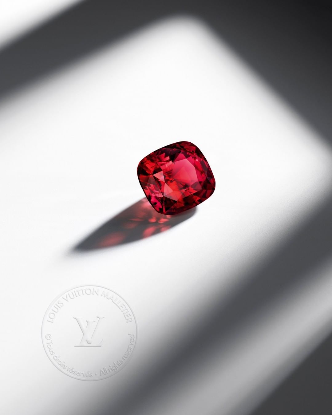 A red gemstone with a cushion cut rests on a white surface with shadows, highlighting its facets and rich color under studio lighting.