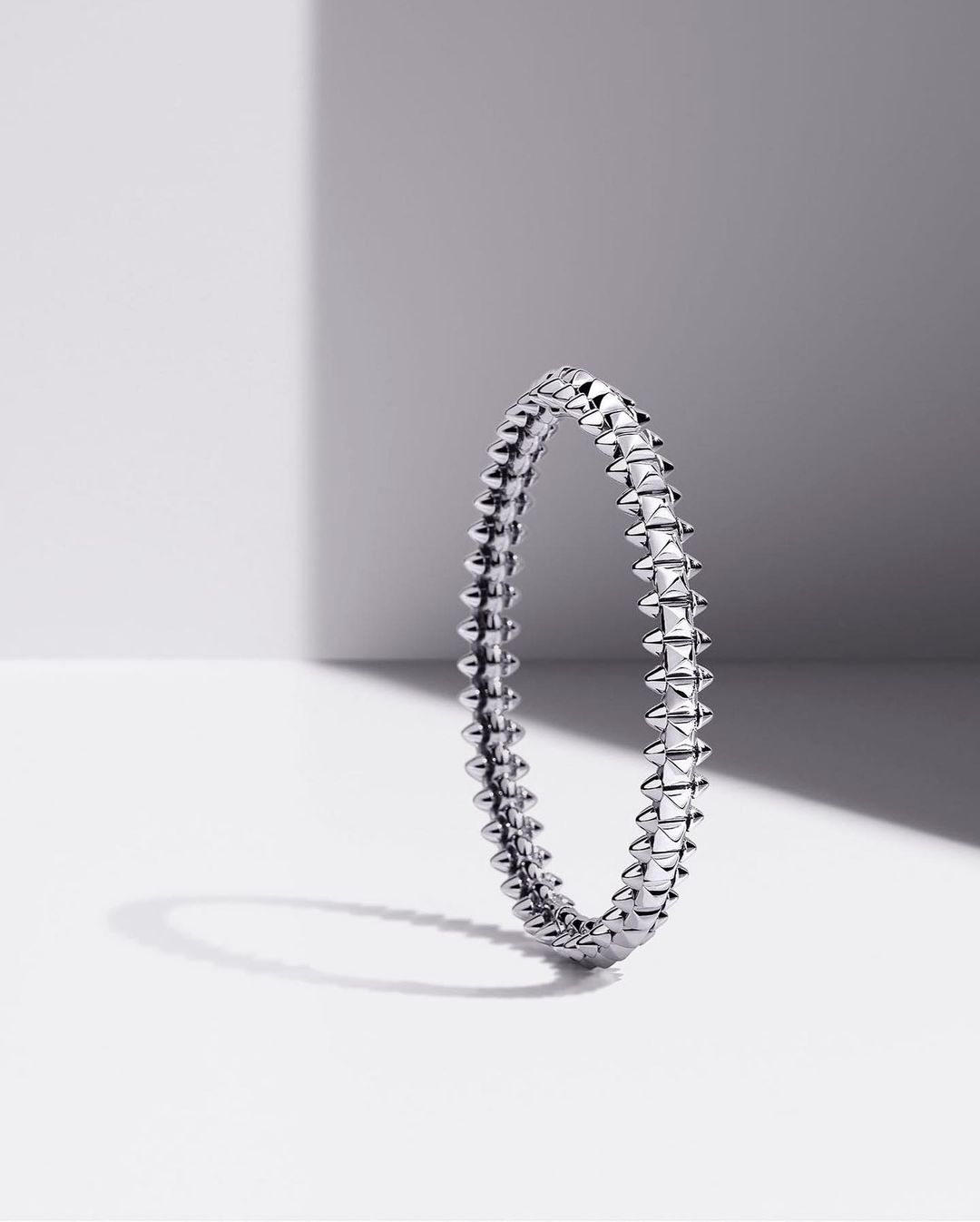 Studded silver bangle stands upright on a white surface with a grey and white background shadow.
