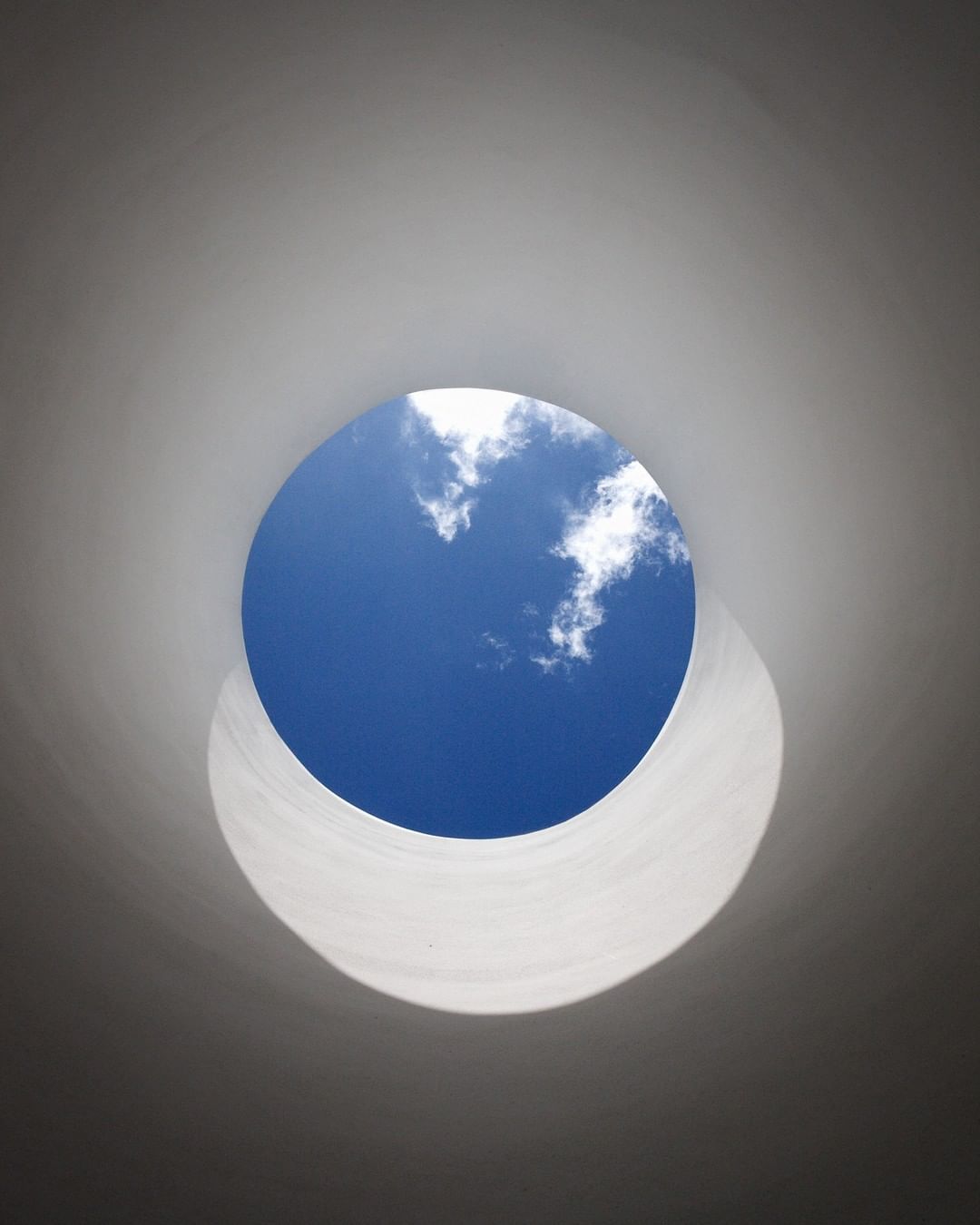 Looking up through a round opening to a blue sky with white clouds. The opening is framed by a white structure, possibly a tunnel or skylight.