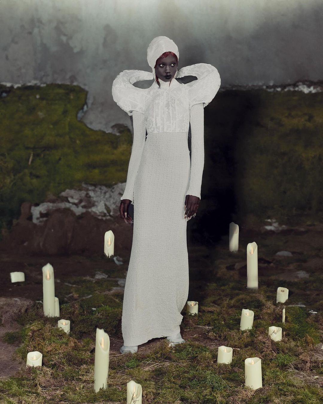 A dark-skinned woman wearing an ornate white dress stands among candles in an outdoor setting with mossy walls in the background.