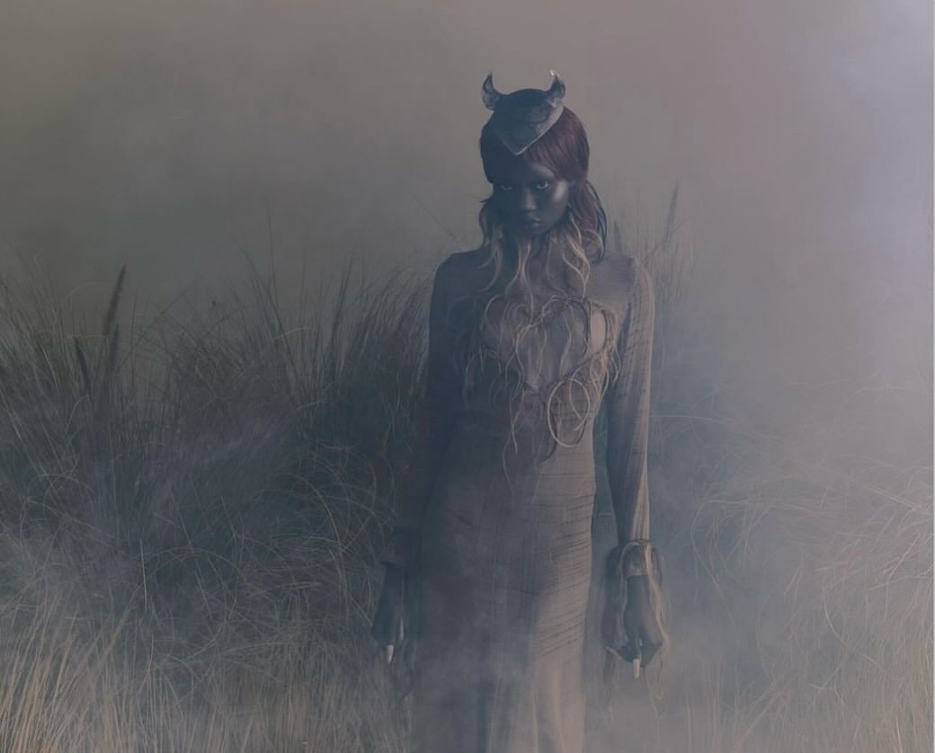 A woman in a costume with small horns poses in a field obscured by smoke and tall grass, with dramatic lighting and muted colours.