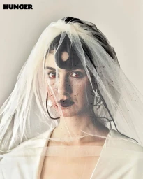 Portrait of a woman wearing a sheer white veil and white dress with dark lipstick and splatters on her face.