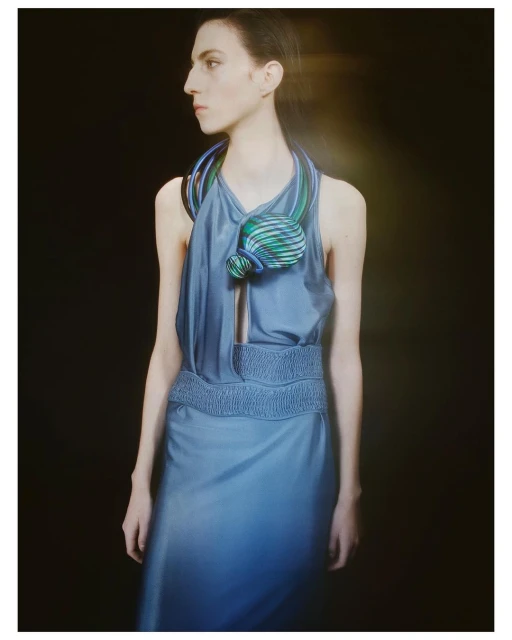 Woman in blue halter dress poses with a decorative neck piece against a dark backdrop in a studio fashion shot.