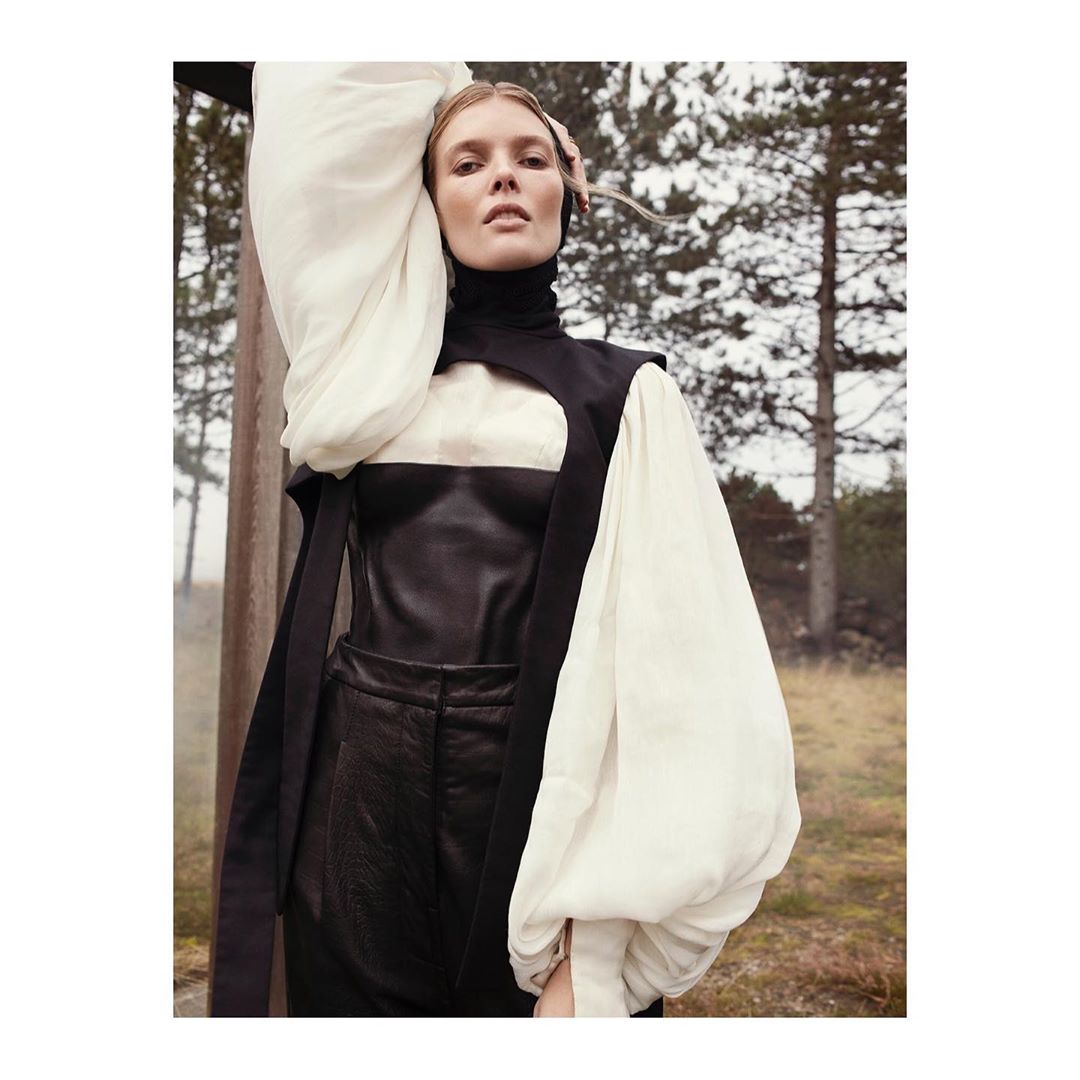 Fashion editorial shot of a blonde woman in a black and white outfit posing in a field with trees in the background, portrait orientation.