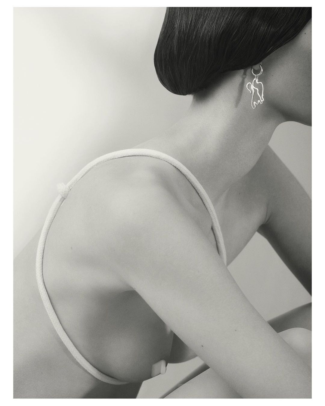 Monochrome portrait of a woman wearing a sculptural silver earring and a thin strap garment against a plain light background.