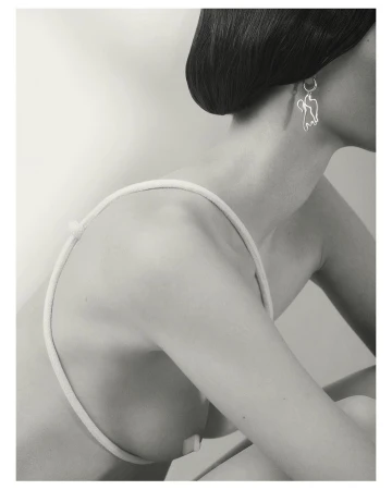 Monochrome portrait of a woman wearing a sculptural silver earring and a thin strap garment against a plain light background.