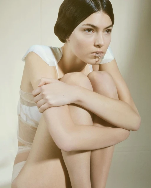 An adult female model poses indoors with her arms around her legs, wearing a white garment and dark hair. The image has a neutral background.