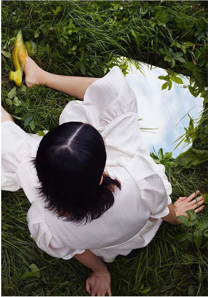 Overhead shot of a woman in a white dress sitting on green grass near a mirror reflecting the sky, showcasing nature and fashion.