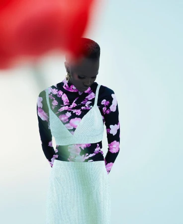 Model with short dark hair wearing a white slip dress over a black floral top against a plain light background.