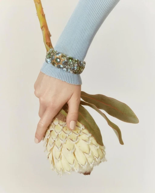 Close-up shot of a female hand wearing a beaded bracelet, delicately holding a large white flower against a plain background.