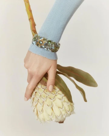 Close-up of a hand wearing a jewel-encrusted bracelet and holding a protea flower against a white background