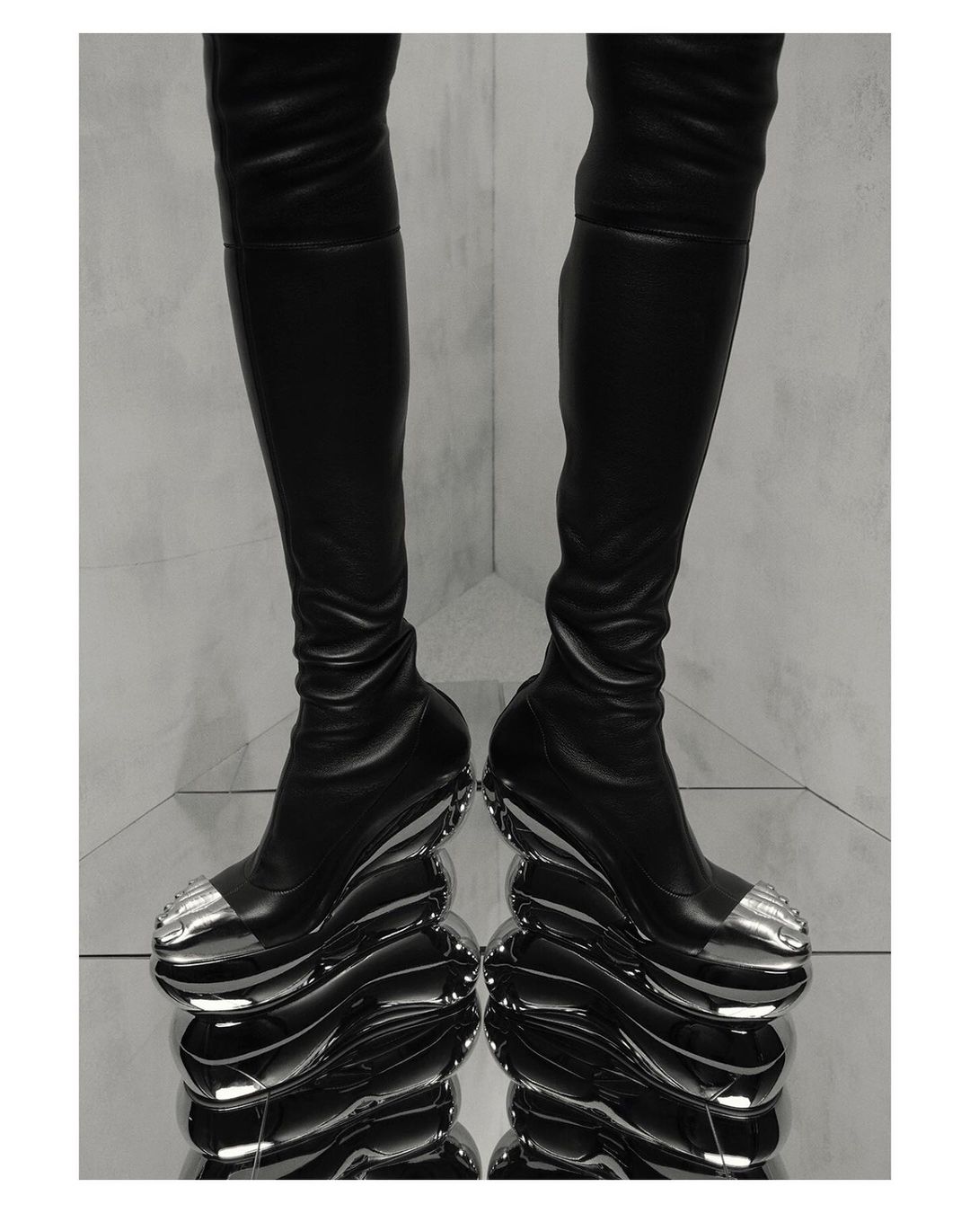 A pair of thigh-high, black leather boots with bulbous, metallic silver platform soles and toe caps, displayed on a mirrored surface.
