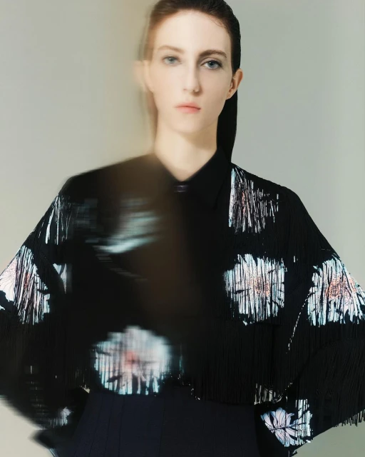 Fashionable young adult woman with fair skin posing in a black top with shimmering floral pattern against a neutral studio backdrop.