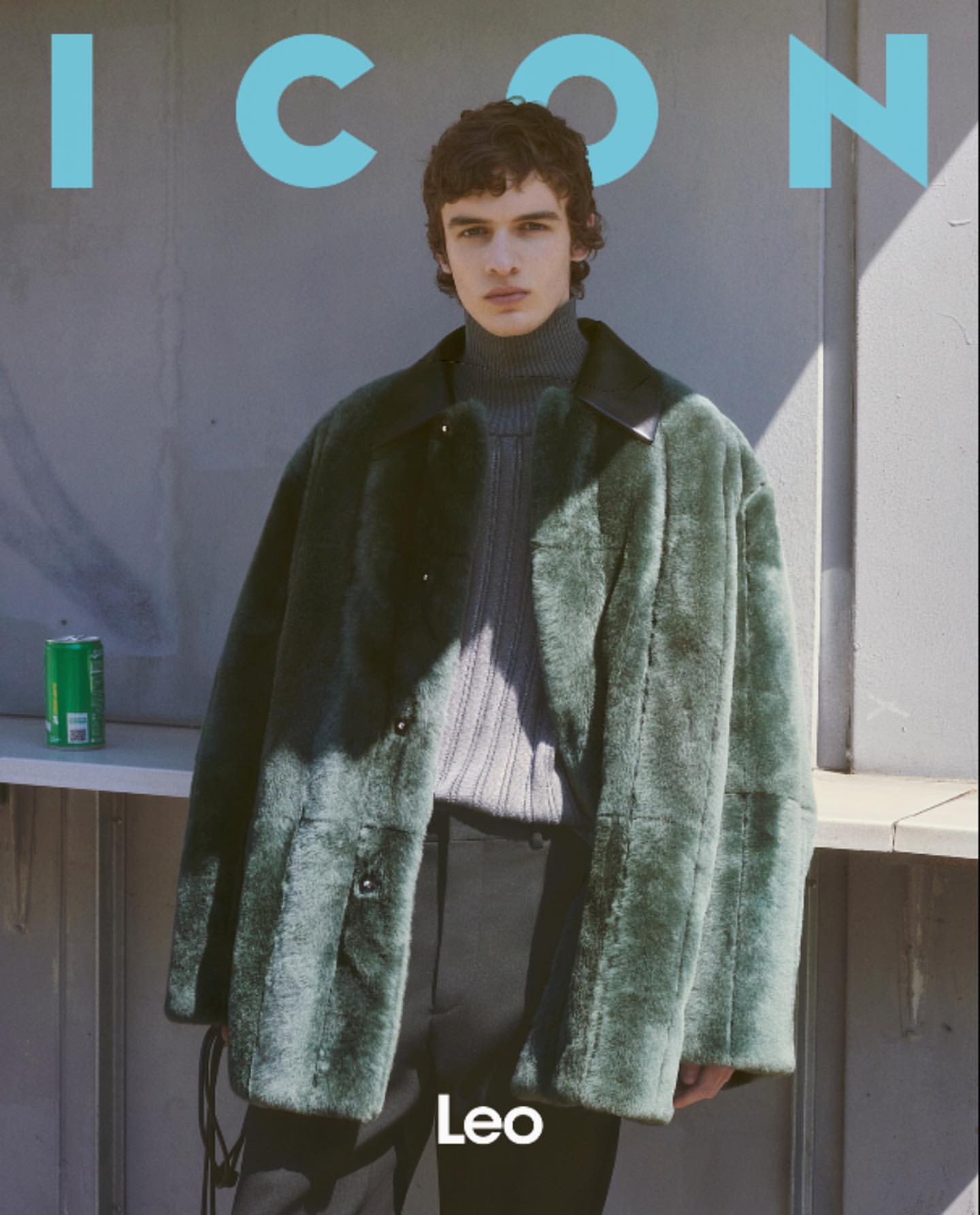 Young man in a green faux fur coat and ribbed grey turtleneck standing against an outdoor wall.