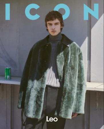Young man in a green faux fur coat and ribbed grey turtleneck standing against an outdoor wall.