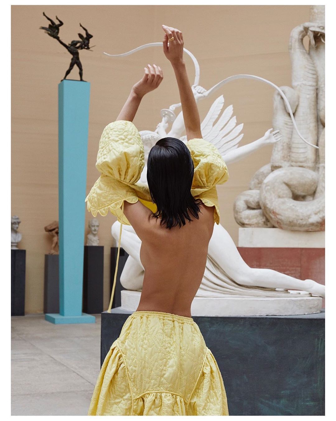 Woman in yellow dress with raised arms poses in an art museum in front of sculptures. Fashion editorial shot.