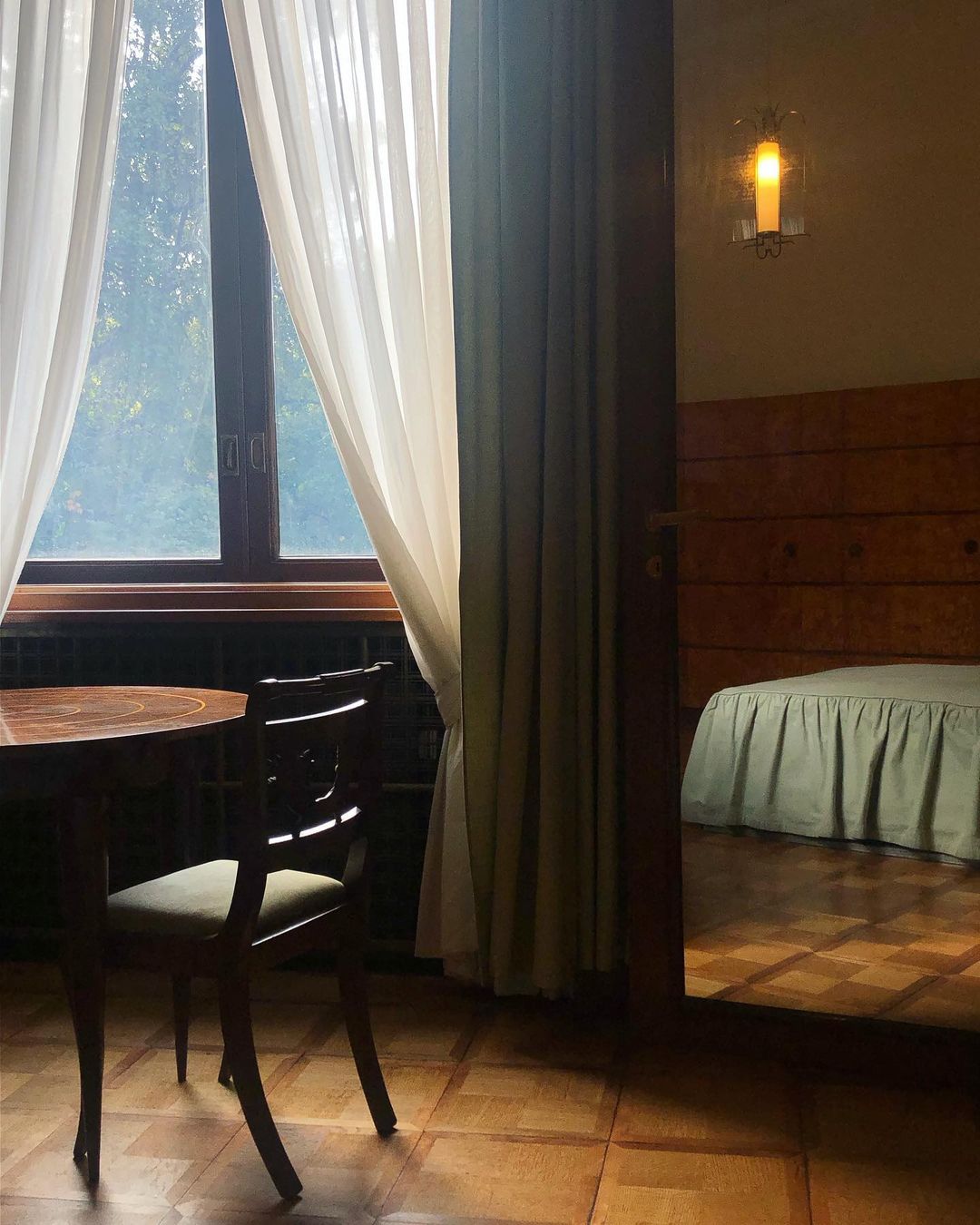 Interior view of a room with a round table, chair, window with curtains, and a glimpse into a bedroom with a bed and parquet floor.