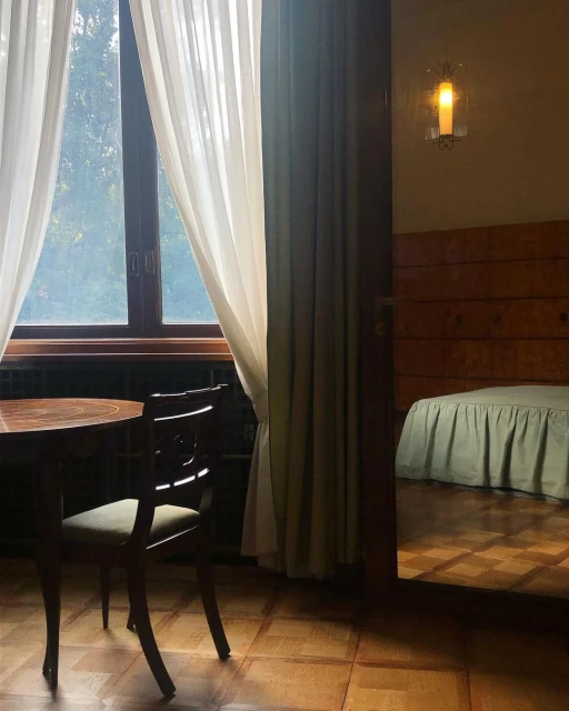 Interior view of a room with a round table, chair, window with curtains, and a glimpse into a bedroom with a bed and parquet floor.
