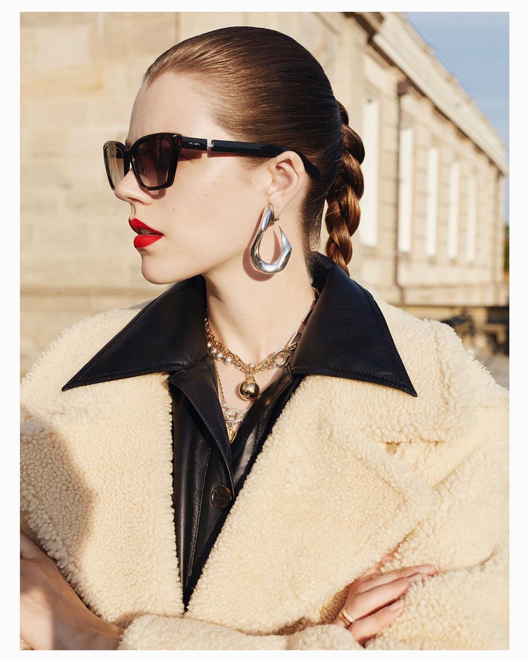 Portrait of a woman with braided hair, wearing a textured beige jacket, oversized earrings, and sunglasses, standing outdoors.