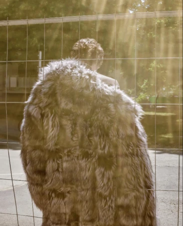 Rear view of a person wearing a fluffy textured faux fur coat behind a wire mesh fence in sunlight by Lea Nielsen.