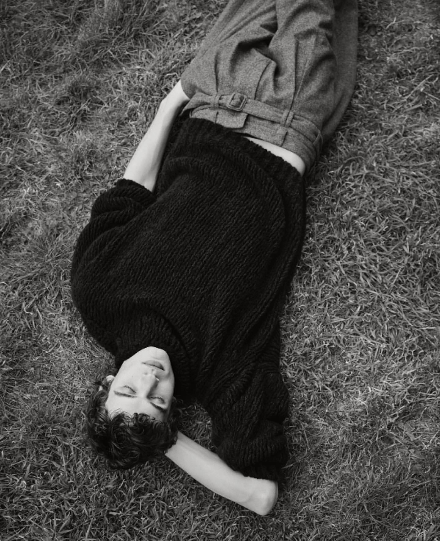 Monochrome portrait of a person lying on grass wearing a black knit sweater and grey belted trousers.
