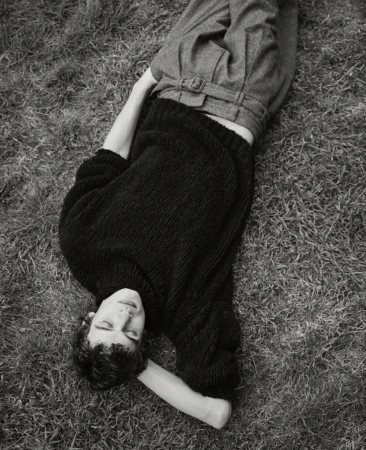 Monochrome portrait of a person lying on grass wearing a black knit sweater and grey belted trousers.