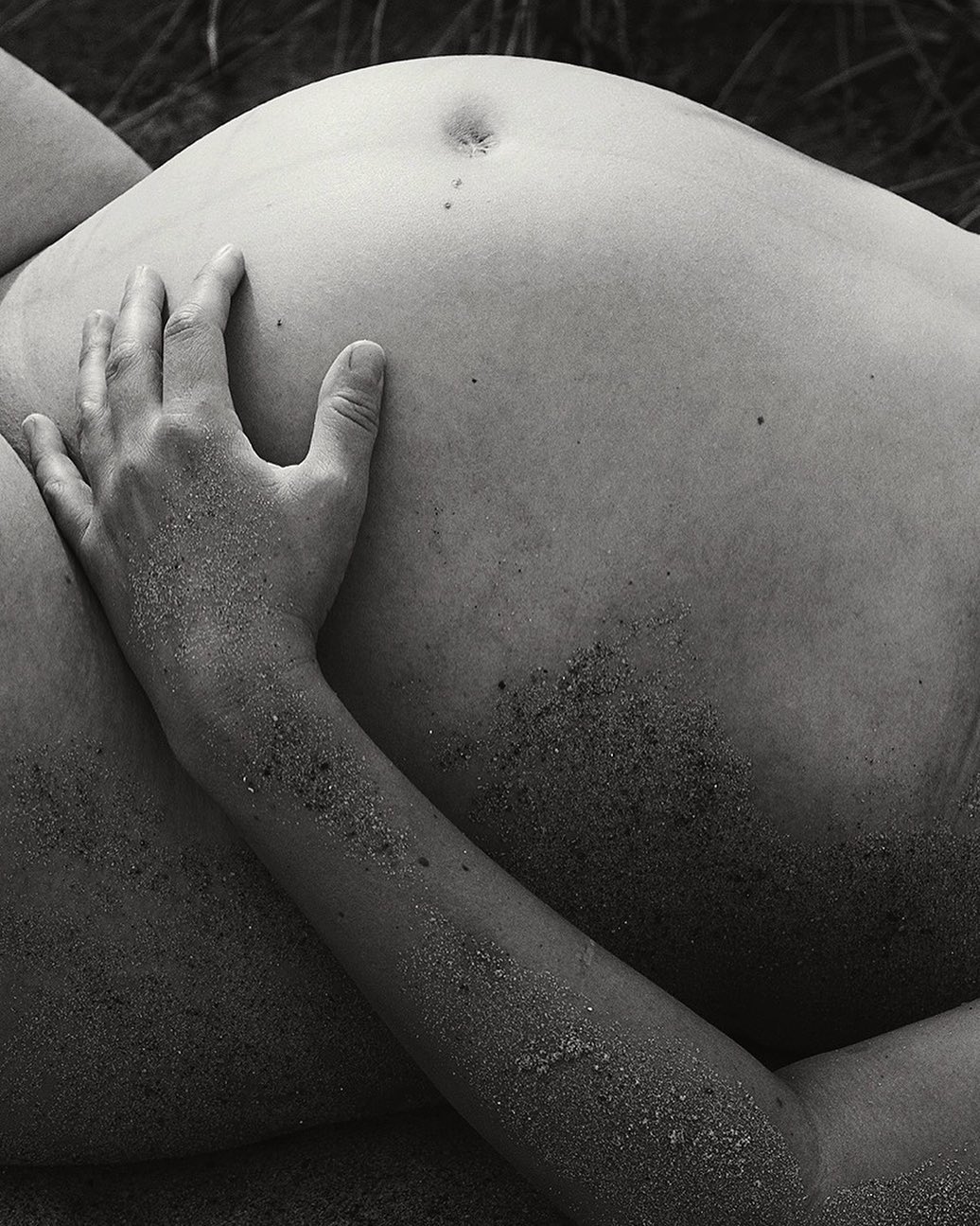 Close-up black and white photograph of a pregnant person's abdomen and hand covered with sand, outdoors against a natural background.