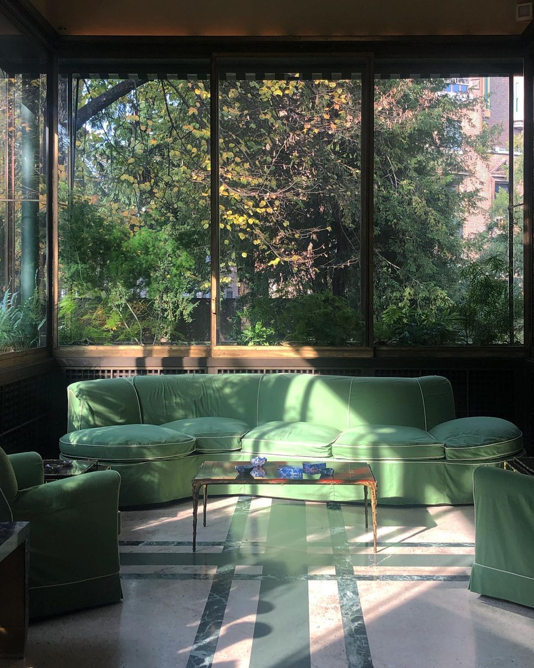 Sunlit interior with green sofa and chairs in a room with large windows looking out onto a green, leafy outdoor scene.
