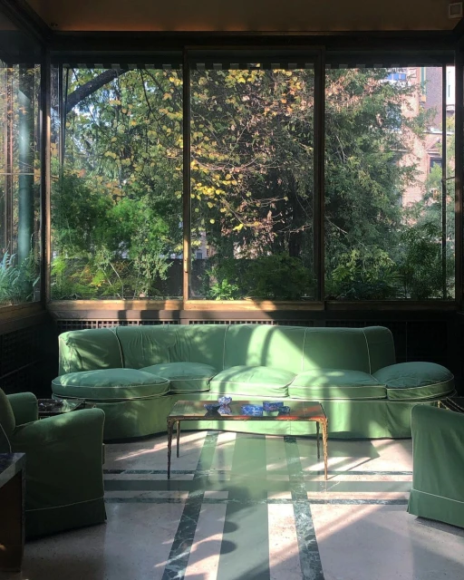 Sunlit interior with green sofa and chairs in a room with large windows looking out onto a green, leafy outdoor scene.