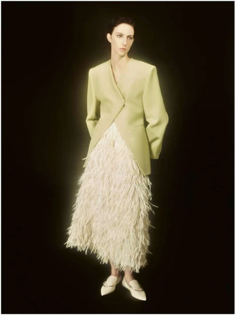 Elegant woman in a light green blazer and textured white skirt poses in a studio with dark background, showcasing fashion-forward style.