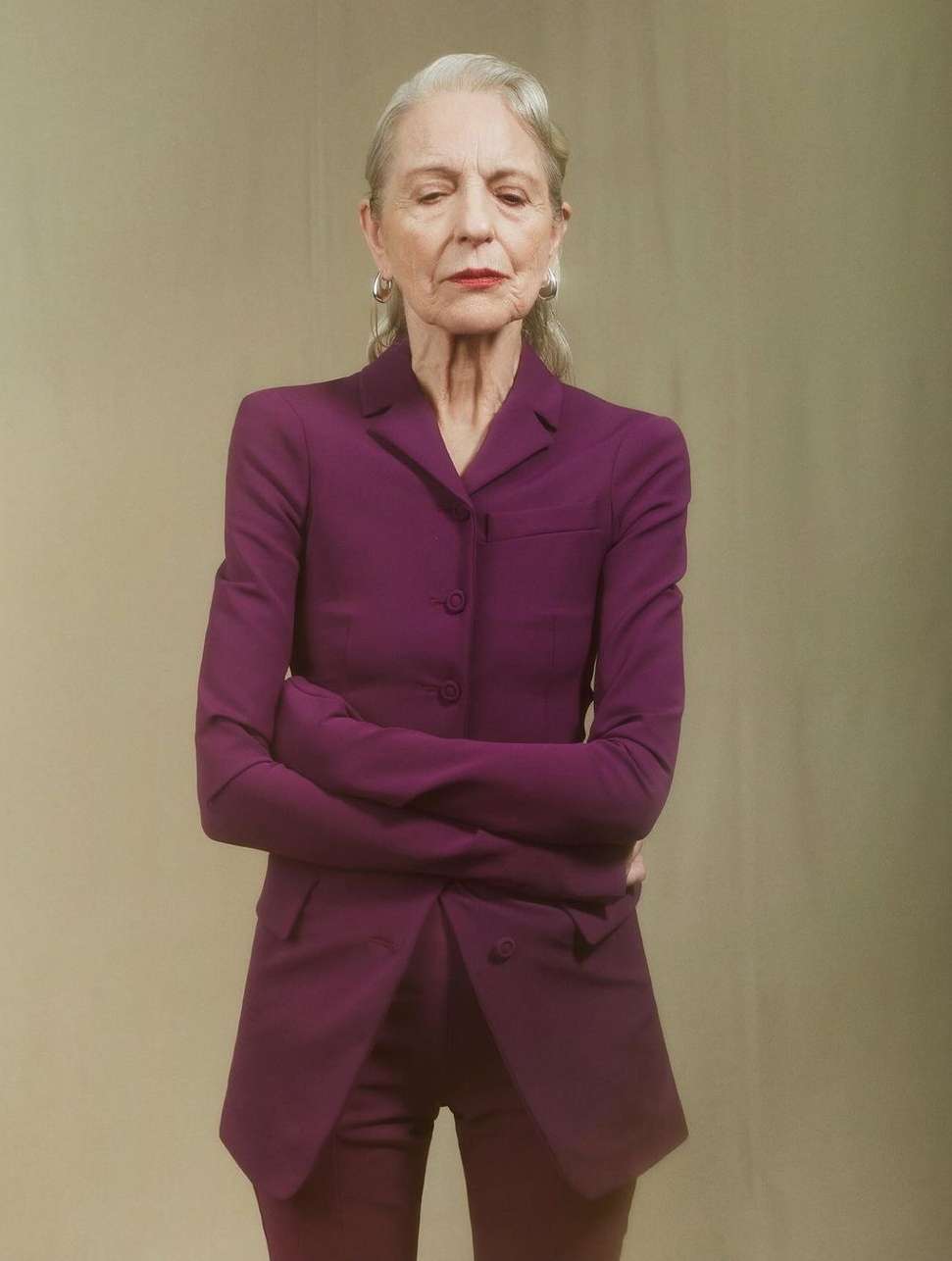 Elegant mature woman in a magenta suit with arms crossed, standing against a neutral background in a professional portrait.
