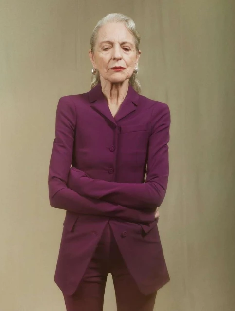Elegant mature woman in a magenta suit with arms crossed, standing against a neutral background in a professional portrait.