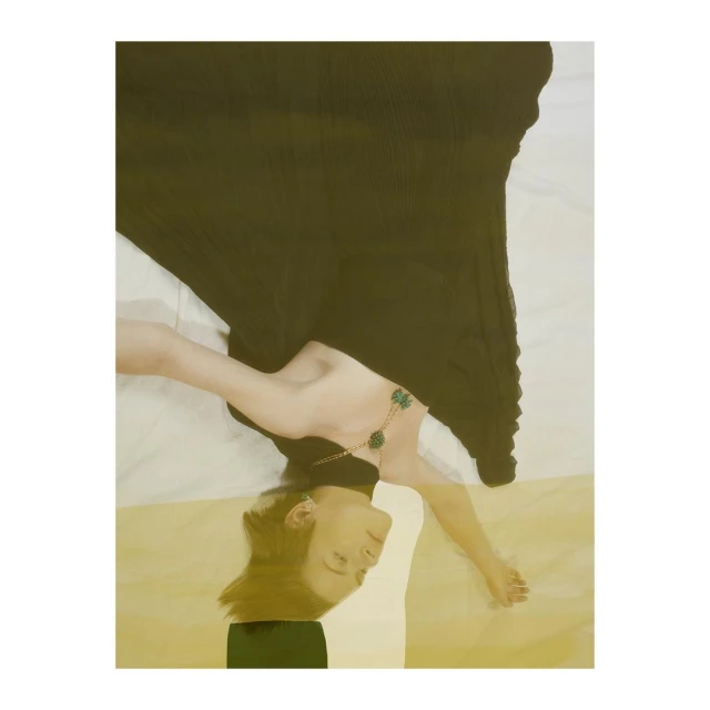 Woman lying down wearing a black dress and green necklace against a white and gold patterned background in a portrait orientation.