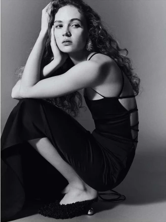 Woman with long curly hair wearing a black slip dress squatting against a grey studio backdrop
