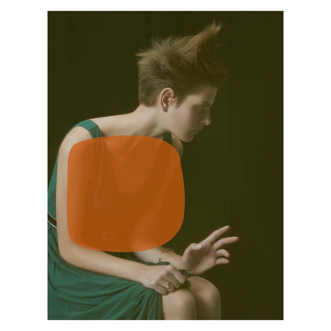 Indoor portrait of a person in a green dress, seated with hands gesturing against a dark background and stylized hair.