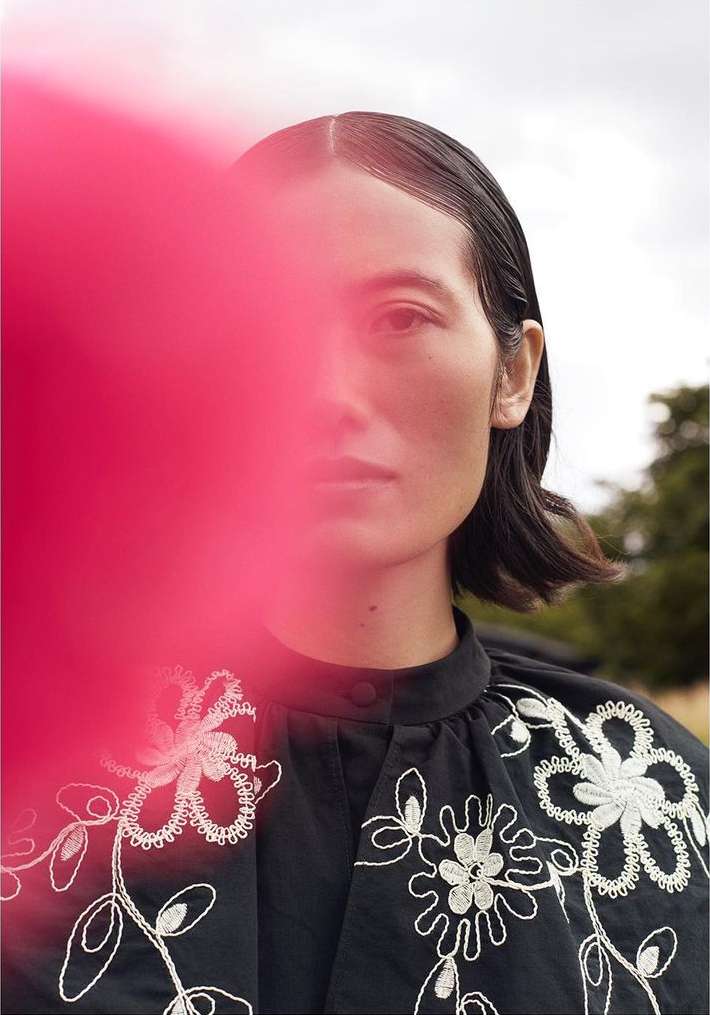 Portrait of a woman with dark hair, wearing a dark blouse with floral embroidery, posing outdoors with pink color cast.