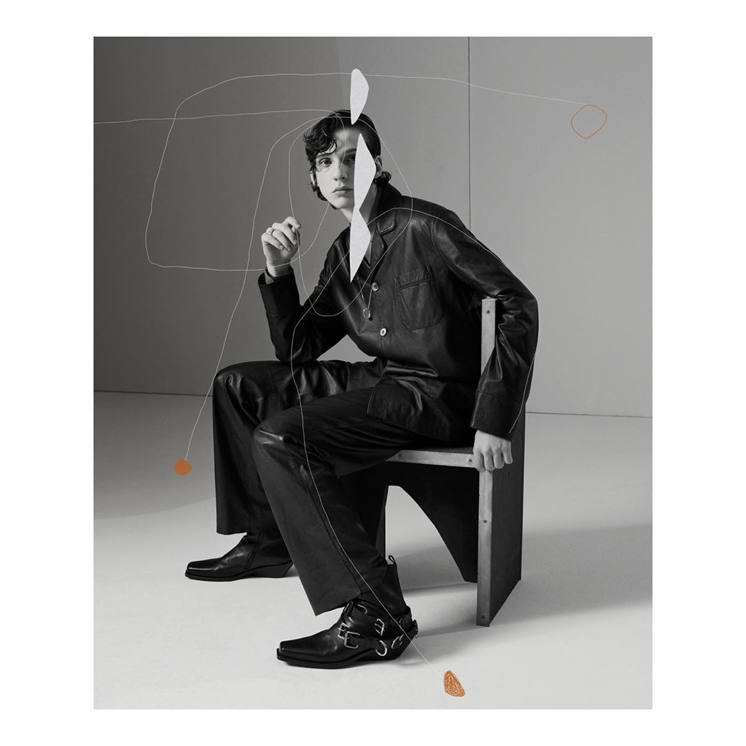 A young adult male model in a dark leather jacket and trousers sits on a chair in a photo studio against a grey backdrop with graphics.