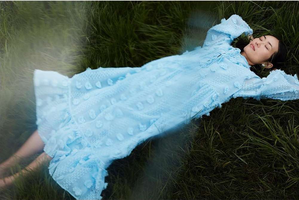 A serene woman in a light blue dress reclines in a field of green grass, eyes closed in relaxation, bathed in soft, diffused light.